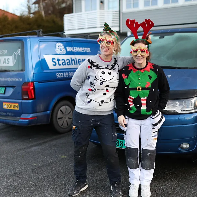 Janne og Christina i julegensere og morsomme briller står foran to blå varebiler fra Staahlen AS.