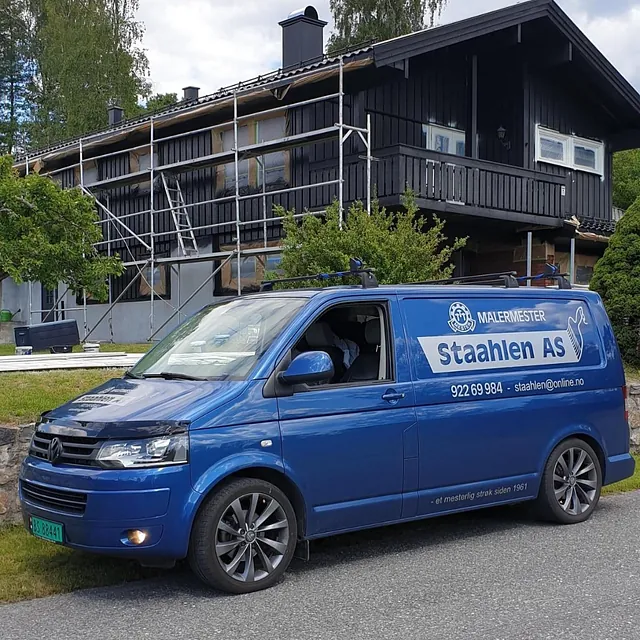 Blå varebil fra Staahlen AS parkert foran et stort hus med stillas og fasadeoppussing pågår.