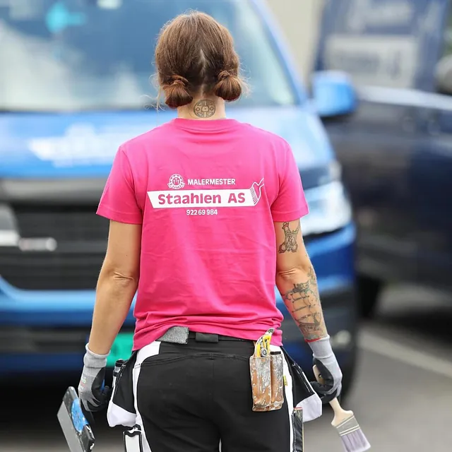 Maler i rosa Staahlen AS-t-skjorte og arbeidsklær sett bakfra, med pensel og sparkelspade i hendene.